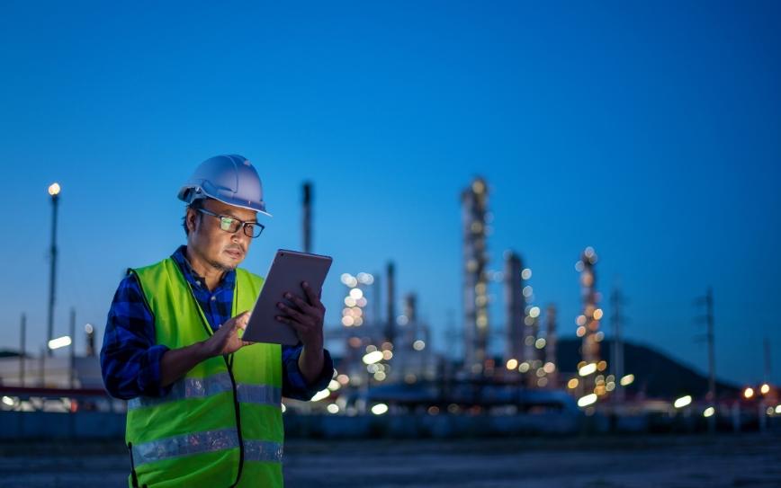 A person wearing a safety vest in an industrial business environment and hard hat looks at a tablet