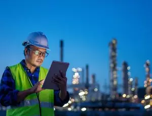 A person reviews a tablet with an industrial facility in the background