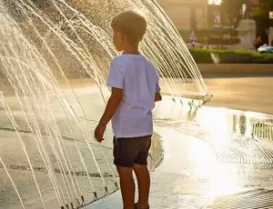 A young child stands by a public water park fountain